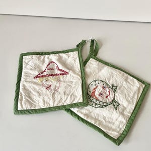 May include: Two white cotton pot holders with green trim. One pot holder has an embroidered image of a person wearing a red hat and the other has an embroidered image of a person wearing a blue and white hat.