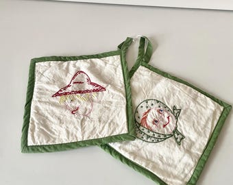May include: Two white cotton pot holders with green trim. One pot holder has an embroidered image of a person wearing a red hat and the other has an embroidered image of a person wearing a blue and white hat.