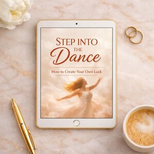 Peut inclure: Une tablette affichant la couverture du livre intitulée Step Into The Dance: How to Create Your Own Luck, illustrant une personne en robe blanche dansant parmi les nuages, accompagnée d'un stylo doré et de deux anneaux dorés à proximité.