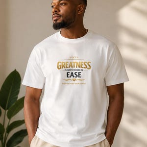 Peut inclure: Un t-shirt blanc à manches courtes avec un texte doré et noir indiquant : FOCUS GREATNESS IS NOT FOUND IN EASE PUSH BEYOND YOUR LIMITS. Le motif comprend un petit emblème doré entre les mots EASE et PUSH BEYOND YOUR LIMITS.