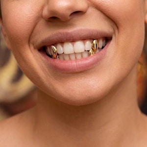 May include: A set of four individual gold tooth caps, known as fangs, placed on the upper and lower canine teeth. The metallic gold covers are smooth and polished, fitted over the natural teeth to create a distinct, reflective, and shiny appearance.