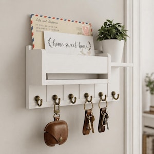 Könnte beinhalten: Weißer Wandorganizer aus Holz mit einem Regal, einem Postschlitz und Schlüsselhaken. Enthält eine kleine Topfpflanze, einen Umschlag und eine Karte mit der Aufschrift "home sweet home". Mehrere Schlüssel und ein braunes Etui hängen an den Haken.