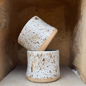 Op de afbeelding: Twee keramische bekers met een gespikkelde witte en bruine afwerking. De onderste beker staat rechtop, terwijl de bovenste beker schuin op de rand balanceert. Beide bekers hebben een gladde, ongeglazuurde beige basis en een textuur.