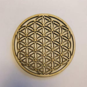 Peut inclure: Un dessous de verre rond en bois clair avec un motif de Fleur de Vie découpé au laser. Le motif géométrique complexe est mis en évidence par un contour brun foncé.