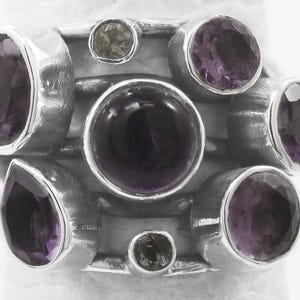 Könnte beinhalten: Ein Sterling Silber Ring mit einer Ansammlung von Edelsteinen. Der Ring zeigt einen großen, dunkelvioletten zentralen Stein, umgeben von kleineren, runden und tropfenförmigen Amethysten und olivgrünen Steinen. Das Design ist modern und auffällig.
