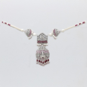 May include: A silver-toned necklace featuring a central pendant with intricate embossed figures, accented by deep red stones and small white pearls. The chain consists of white beads and red stone segments, creating a symmetrical design.