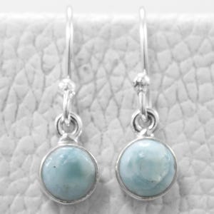 Può includere: Un paio di orecchini pendenti in argento con pietre preziose rotonde azzurro chiaro. Gli orecchini hanno un design semplice con un piccolo gancio e un'incastonatura a castone attorno a ogni pietra. Le pietre hanno un aspetto marmorizzato.