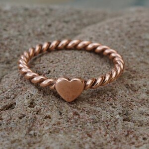 Op de afbeelding: Een roségoudkleurige ring met een gedraaide, touwachtige band en een kleine, gladde hartvormige bedel in het midden. Het metaal heeft een gepolijste afwerking die het licht reflecteert en de textuur van het gedraaide bandontwerp benadrukt.