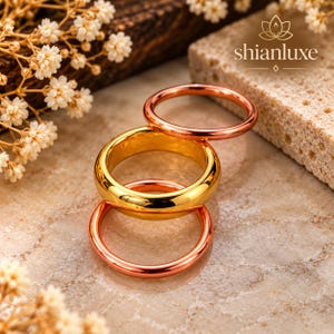 May include: A set of three smooth, polished metal rings stacked together. The set includes one thick gold-toned band positioned in the centre, flanked by two thinner rose gold-toned bands. The brand name shianluxe is printed in the upper right corner.