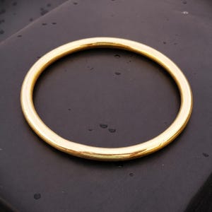 May include: A polished gold-tone bangle bracelet with a smooth, rounded surface and a circular shape. The metal has a reflective finish that catches the light, creating a bright appearance against the dark, matte surface underneath.