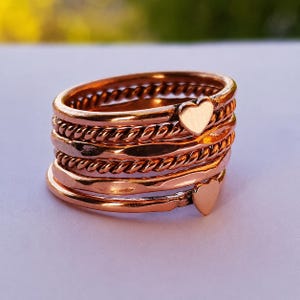 Puede incluir: Un conjunto de cinco anillos de tono cobre con una mezcla de texturas, incluyendo dos bandas de cuerda trenzada, dos bandas lisas y una banda martillada. Dos de los anillos están adornados con pequeños dijes planos en forma de corazón.