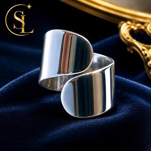 May include: A polished silver-tone bypass ring featuring a wide, smooth, and reflective band with rounded ends that overlap slightly. The metal surface has a high-gloss finish that captures light and reflections, creating a sleek, continuous appearance.