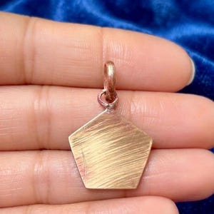 Puede incluir: Un colgante dorado con forma de pentágono. El colgante tiene un lazo en la parte superior para una cadena. La superficie del colgante tiene una textura cepillada. El colgante está en la mano de una persona.