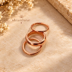 Könnte beinhalten: Drei polierte Ringe aus roségoldfarbenem Metall, die vertikal gestapelt sind. Die Ringe haben ein glattes, abgerundetes Banddesign. Der Text shianluxe ist in Dunkelbraun über den Ringen aufgedruckt, begleitet von einem kleinen Lotusblüten-Symbol.