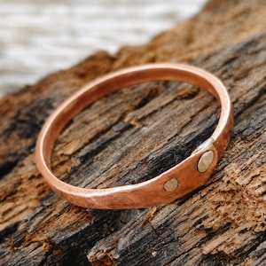 May include: A circular copper bangle bracelet with a hammered texture. The band features a slightly overlapping design secured by two small, round, silver-toned metal rivets positioned near one end of the copper metal loop.