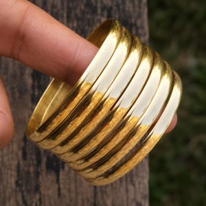 Puede incluir: Un conjunto de seis brazaletes apilados de metal en tono dorado con una superficie lisa, pulida y reflectante. Las pulseras circulares son uniformes en ancho y grosor, presentando un perfil simple y redondeado de aspecto dorado brillante.