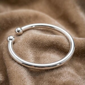 Op de afbeelding: Een glanzende zilveren armband met een afgerond, open ontwerp. De armband heeft een glad, gepolijst oppervlak en kleine bolvormige accenten aan elk open uiteinde. Het sieraad wordt gepresenteerd op een zacht, bruin, getextureerd oppervlak.