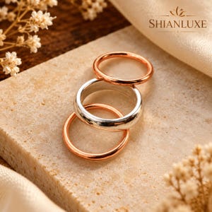 Peut inclure: Un ensemble de trois bagues comprenant deux anneaux fins en or rose et un anneau plus large en argent. Les bagues sont empilées, présentant une finition métallique lisse et polie. Le nom de marque SHIANLUXE est imprimé en haut à droite.