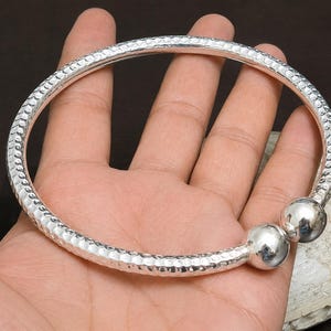 Op de afbeelding: Een zilverkleurige open armband met een getextureerd, gehamerd oppervlak langs de band en twee gladde, gepolijste bolvormige uiteinden aan beide kanten. Het metaal heeft een reflecterende afwerking en een cirkelvormig, open ontwerp.