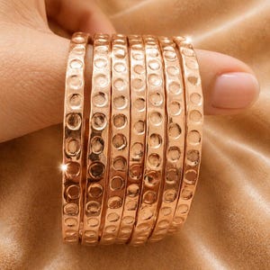 Op de afbeelding: Een stapel van acht koperkleurige armbanden. Elke armband heeft een getextureerd, gehamerd ontwerp met gelijkmatig verdeelde cirkelvormige inkepingen. De sieraden worden gepresenteerd tegen een zachte, neutraal getinte achtergrond.