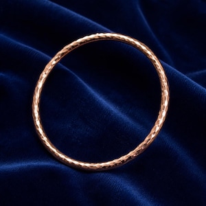 May include: A hammered copper bangle bracelet. The bracelet has a textured surface and a circular shape. The copper colour contrasts with the dark blue velvet background. This piece of jewellery is suitable for everyday wear.
