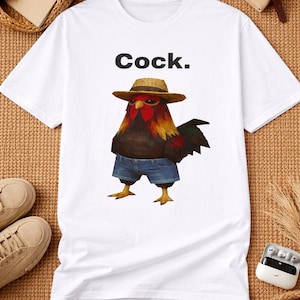 Op de afbeelding: Wit T-shirt met een cartoon haan met een strohoed en een spijkerbroek. Het woord "Cock." staat boven de haan gedrukt. De haan heeft rode, bruine en gele veren. Het T-shirt ligt op een bruin geweven oppervlak.