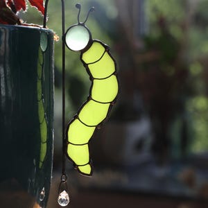 Könnte beinhalten: Ein Buntglas-Ornament in Form einer Raupe in hellem Limettengrün mit einem durchscheinenden weißen Kopf und dunklen Metall-Lötlinien. Die Raupe ist an einem dünnen, gebogenen Metallhaken befestigt, der über den Rand eines dunkelblauen Keramiktopfs hängt.