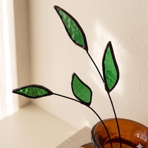 May include: A decorative glass leaf stem with three green leaves and dark brown outlines. The leaves are set against a light background, with the stems emerging from a brown glass vase.