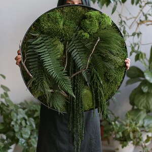 May include: A circular wall art piece featuring a dense arrangement of preserved green moss, various fern fronds, and thin, bare wooden branches. The botanical composition is contained within a thin, black circular frame, creating a textured display.