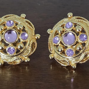 May include: A pair of round gold-tone earrings featuring a central lavender stone surrounded by four smaller lavender stones and gold spheres. The circular frame has a twisted rope texture along the outer edge with small protruding gold beads.