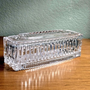 May include: A rectangular clear glass butter dish with a lid. The base and lid feature a vertical ribbed pattern along the sides, creating a textured appearance. The top of the lid has a smooth, flat rectangular surface with beveled edges.