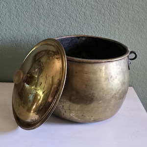 Pode incluir: Uma panela redonda de latão com tampa a condizer, com um pequeno puxador de madeira. A panela tem um corpo largo e profundo e uma pequena pega em anel de metal na lateral. A superfície de latão apresenta sinais de desgaste e pátina.