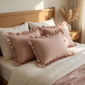 May include: Four blush pink rectangular pillows with ruffled edges rest on a bed with white and pink bedding. The pillows are arranged in a layered composition, creating a cozy and inviting aesthetic. The bed is set against a light wood headboard.