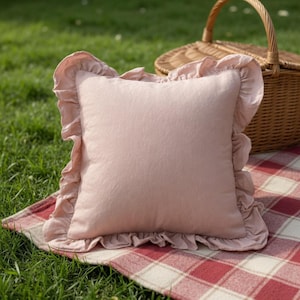 May include: A square, blush-colored pillow with ruffled edges, resting on a red and white checkered blanket. A wicker picnic basket is in the background, set on a grassy lawn. The pillow is approximately 16 inches square.