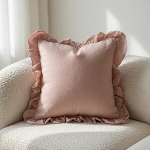 May include: A square, blush-colored pillow with a ruffled edge. The pillow is made of a soft fabric and sits on a cream-colored, textured chair. The ruffled trim adds a decorative element to the pillow.