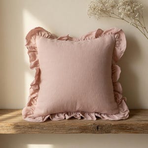 May include: A square, blush-colored pillow with a ruffled edge. The pillow is made of a soft-looking fabric and sits on a rustic wooden surface. A sprig of white flowers is in the background.