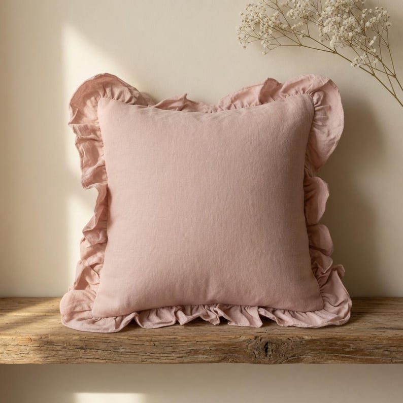 May include: A square, blush-colored pillow with a ruffled edge. The pillow is made of a soft-looking fabric and sits on a rustic wooden surface. A sprig of white flowers is in the background.
