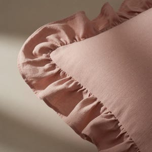 May include: A close-up of a dusty rose-colored pillow with a ruffled edge. The pillow has a soft, textured appearance, and the ruffled trim adds a decorative element. The fabric appears to be linen or a similar material.