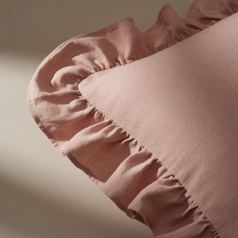 May include: A close-up of a dusty rose-colored pillow with a ruffled edge. The pillow has a soft, textured appearance, and the ruffled trim adds a decorative element. The fabric appears to be linen or a similar material.