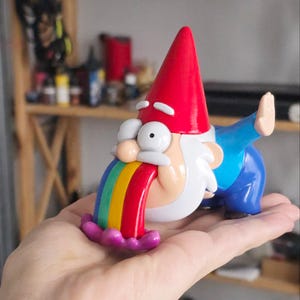 May include: A whimsical gnome figurine with a red hat, white beard, and blue pants. The gnome is depicted vomiting a rainbow. The figurine is held in a hand, showcasing its colorful design and playful theme.