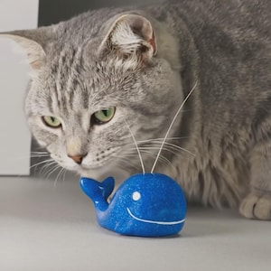 May include: A blue whale-shaped cat toy with a smiling face and white accents. The toy has a glittery finish and two white antennae. A gray cat with green eyes is looking at the toy.