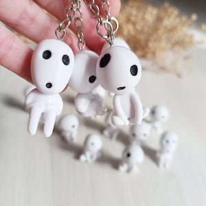 Könnte beinhalten: Weiße Kodama-Geist-Schlüsselanhänger mit schwarzen Augen und kleinen Mündern. Die Schlüsselanhänger sind an silbernen Ketten befestigt. Mehrere kleinere Kodama-Figuren sind darunter verstreut. Die Kodama sind eine Figur aus dem Zeichentrickfilm Prinzessin Mononoke.