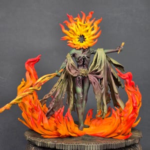 May include: A painted miniature figurine of a humanoid creature with a flaming head, standing amidst sculpted orange and yellow flames. The figure has a dark, textured body draped in tattered, muted green fabric and holds a thin, glowing orange rod.