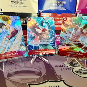 May include: Three One Piece trading cards displayed on clear plastic stands. The left card features Monkey D. Luffy, the center card shows Edward Newgate, and the right card depicts Portgas D. Ace. Each card has vibrant, holographic artwork and text.