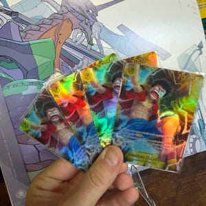 May include: Four holographic trading cards featuring a character in a red shirt and blue pants. The cards have a rainbow sheen and the number "7000" is visible. The background includes a colorful illustration and a clear card stand.