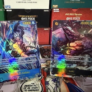 May include: Two One Piece trading cards displayed on stands. The left card features Rocks D. Xebec with a cost of 10 and 12000 power. The right card features Loki with a cost of 6 and 8000 power. Both cards have holographic foil details.