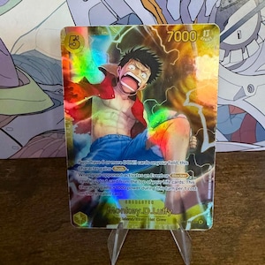 May include: A holographic trading card featuring Monkey D. Luffy from the One Piece series. The card displays an illustration of Luffy with a dynamic pose, wearing a red jacket and blue pants. The card has a power level of 7000 and includes game text.
