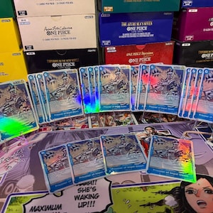May include: A collection of One Piece trading cards featuring the character Buggy, labeled as Prisoner of Impel Down. The cards have a blue border, a power level of 6000, and a holographic finish. They are displayed in front of several stacked One Piece card game boxes.