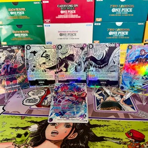 May include: Five One Piece trading cards displayed on clear stands, featuring colorful character illustrations and text. The cards include Saint Gunko, Saint Shamvock, and three cards labeled MMA. The background shows stacked One Piece card game boxes.