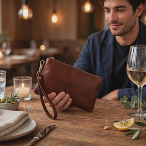 May include: A brown leather wristlet clutch featuring a top zip closure and a detachable wrist strap with a gold-toned metal clasp. The rectangular pouch has a smooth, matte finish and is held in a hand, showcasing its compact and functional design.